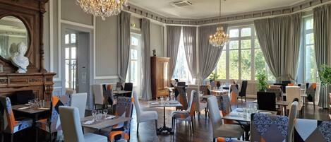 A spacious dining area with multiple tables and chairs, large windows with curtains, and a chandelier.