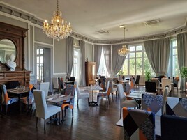 Uma ampla sala de jantar com várias mesas e cadeiras, janelas grandes com cortinas e um lustre.