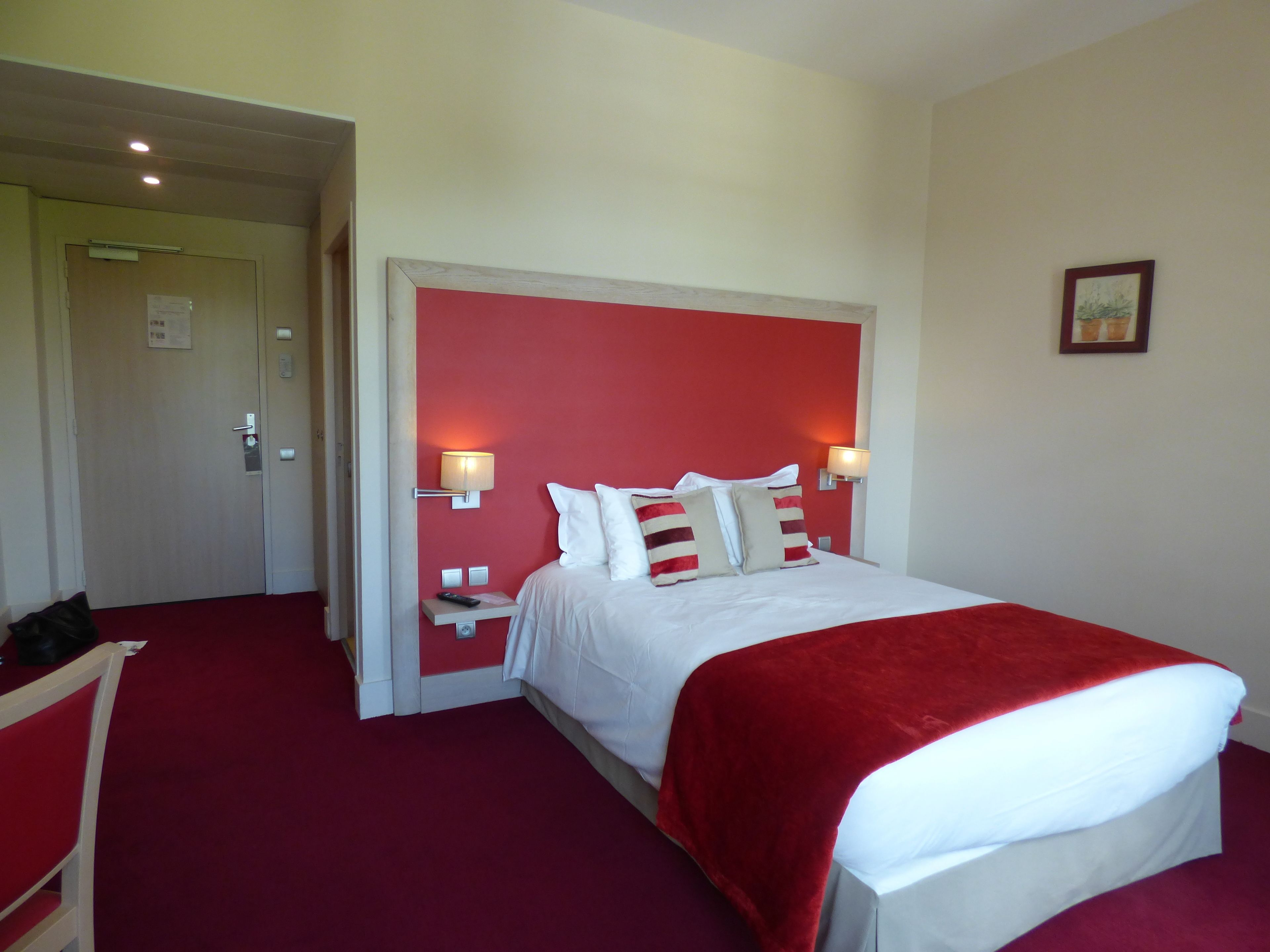 Habitación de hotel con una cama grande, alfombra roja, dos lámparas de noche y una puerta que da a otra habitación.