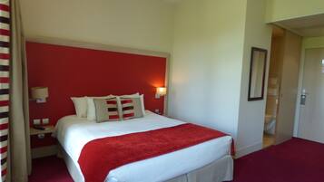 A hotel room with a large bed, a red accent wall, a bedside lamp, and a door leading to a bathroom.