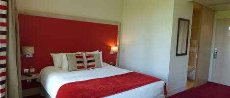 A hotel room with a large bed, a red accent wall, a bedside lamp, and a door leading to a bathroom.