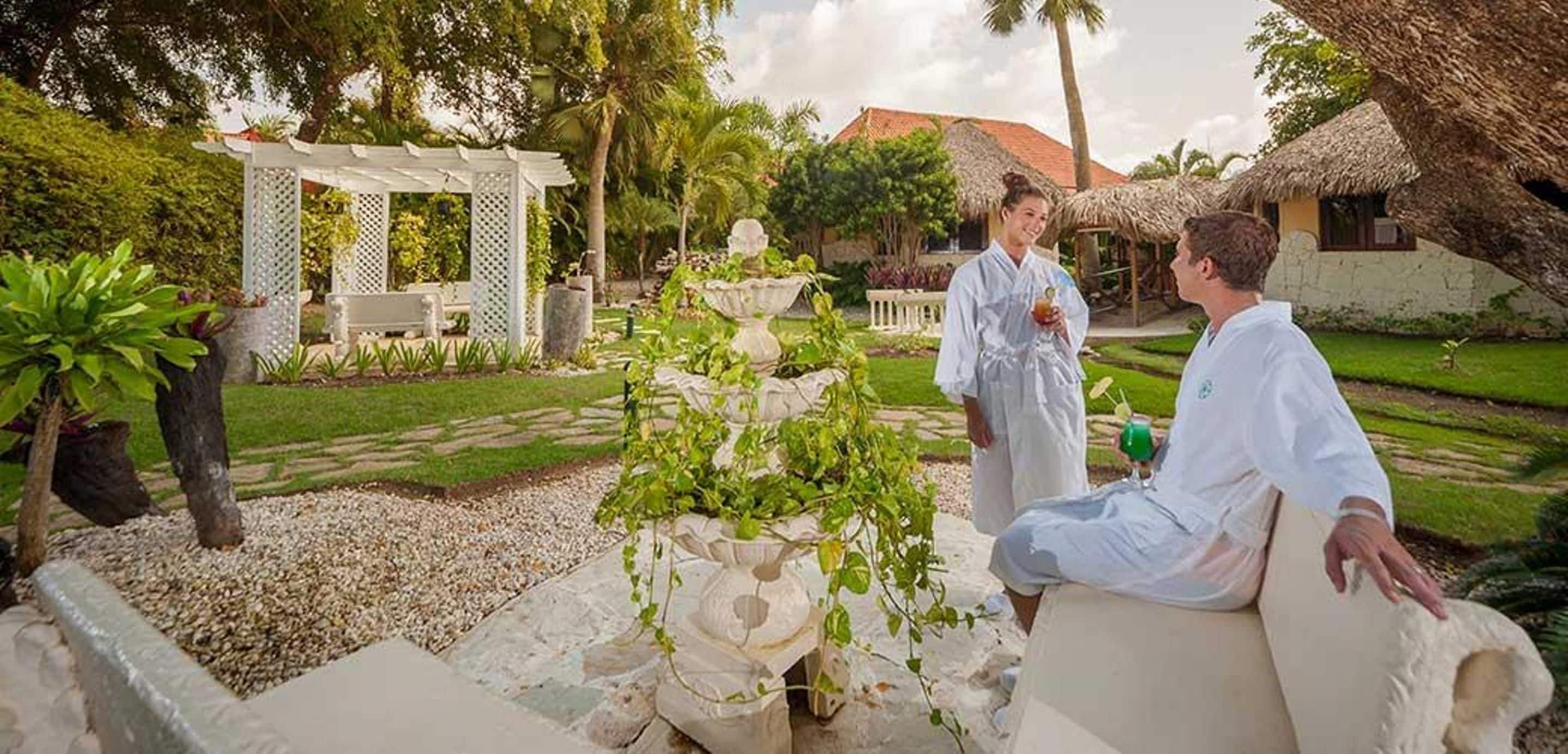 A garden with a white gazebo, a stone fountain, and a thatched-roof building.