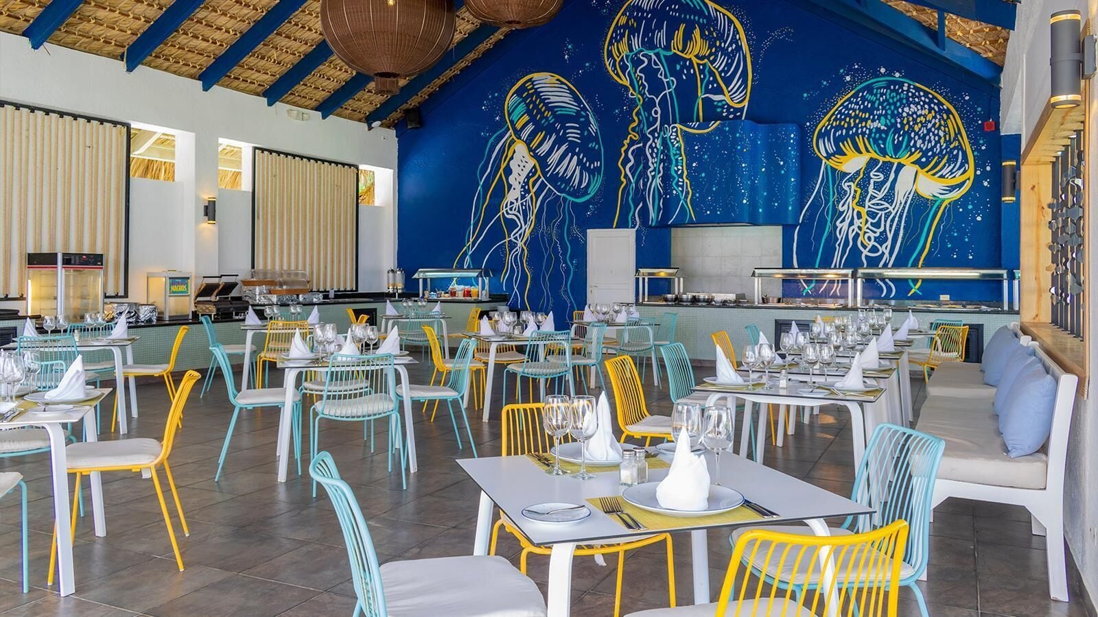 A restaurant with colorful chairs and tables, a blue ceiling, and a mural of jellyfish on the wall.