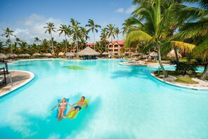 A large swimming pool with two people on inflatable floats, surrounded by palm trees and a resort building.