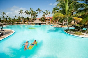 A large swimming pool with two people on inflatable floats, surrounded by palm trees and a resort building.