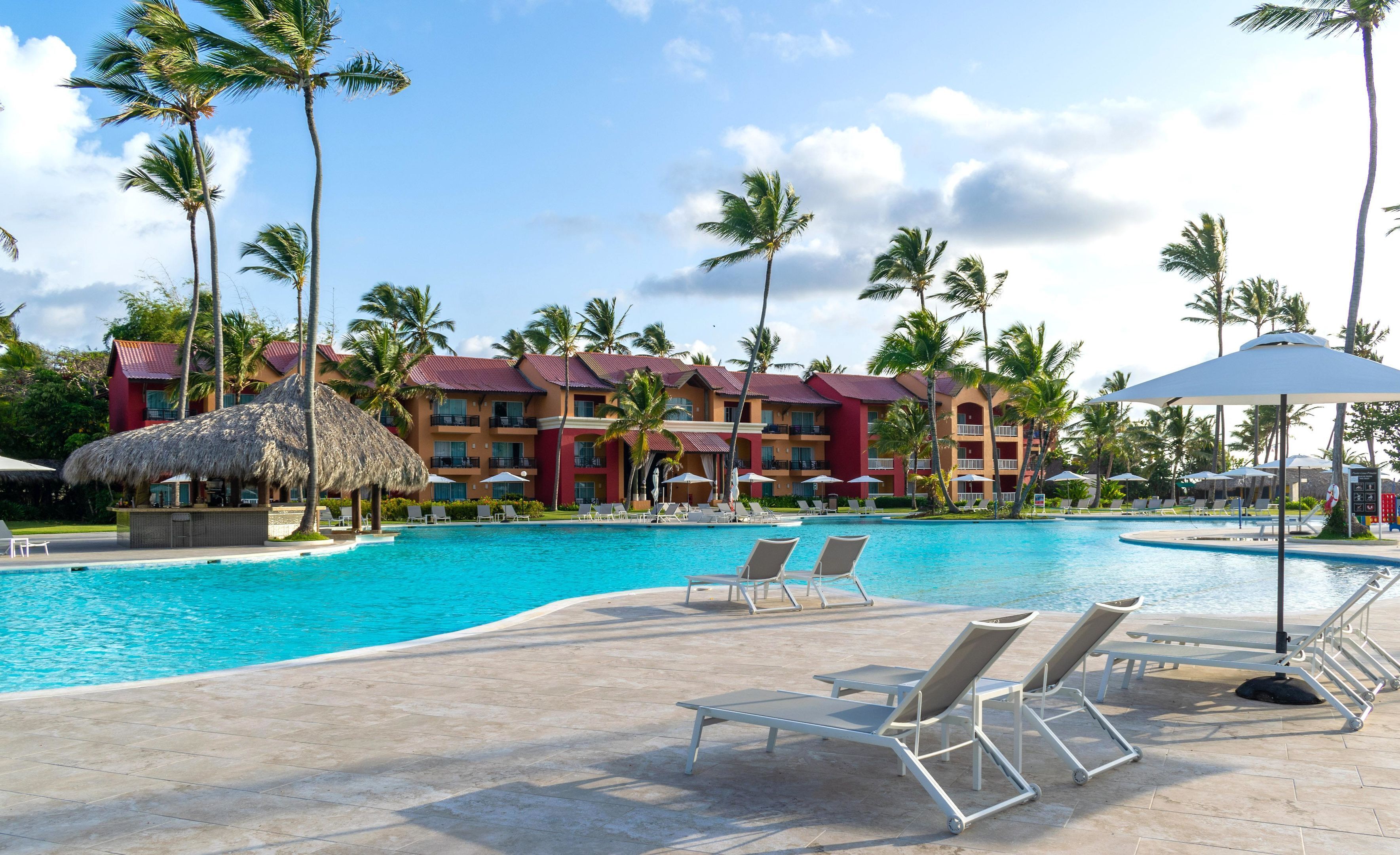 A resort pool area with lounge chairs, palm trees, and a building with multiple balconies.