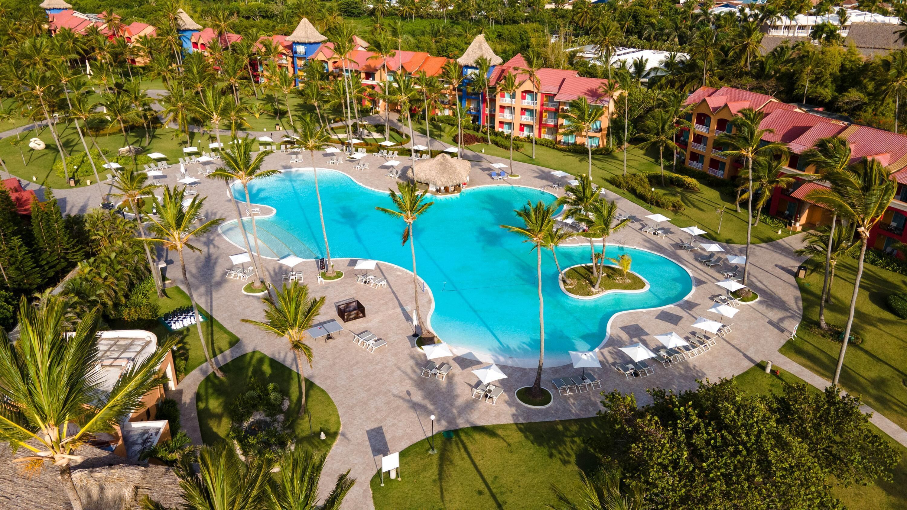 An aerial view of a resort with a large central pool, multiple buildings with red roofs, and surrounding palm trees.