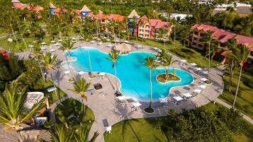An aerial view of a resort with a large central pool, multiple buildings with red roofs, and surrounding palm trees.