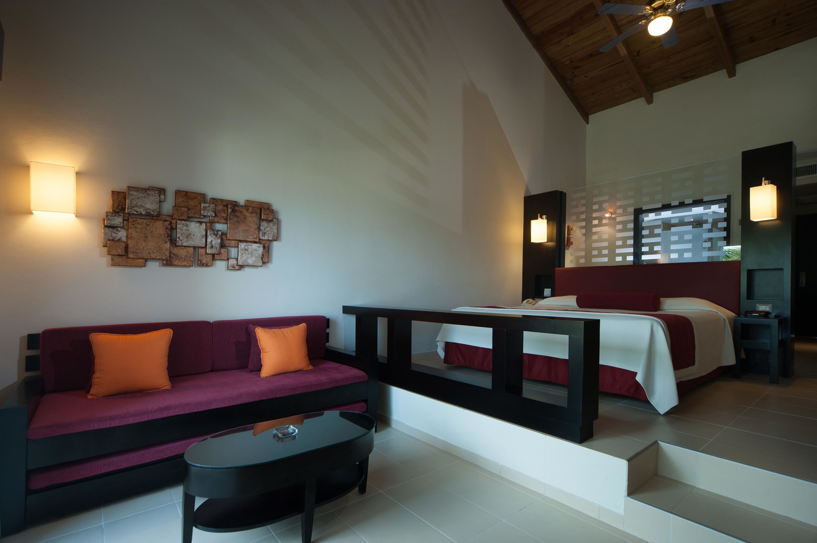 A modern hotel room with a sofa, a small table, a bed with a red and white bedspread, and a wall-mounted light fixture.