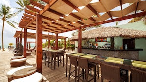 Une terrasse en bois avec des meubles en osier, un coin bar doté d  ’un toit de chaume et, au fond, des palmiers.