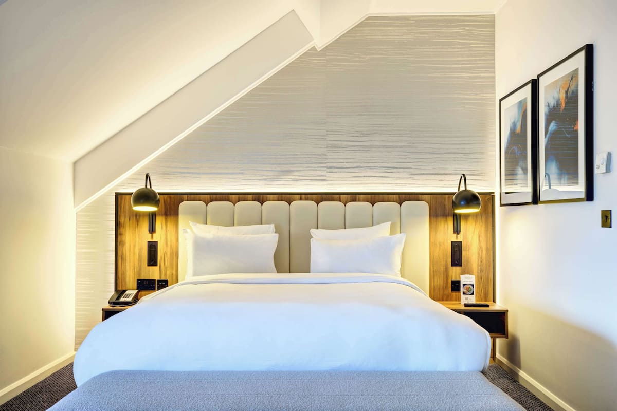 Une chambre d’hôtel avec un grand lit, une tête de lit en bois, deux lampes de chevet et un tableau encadré au mur.