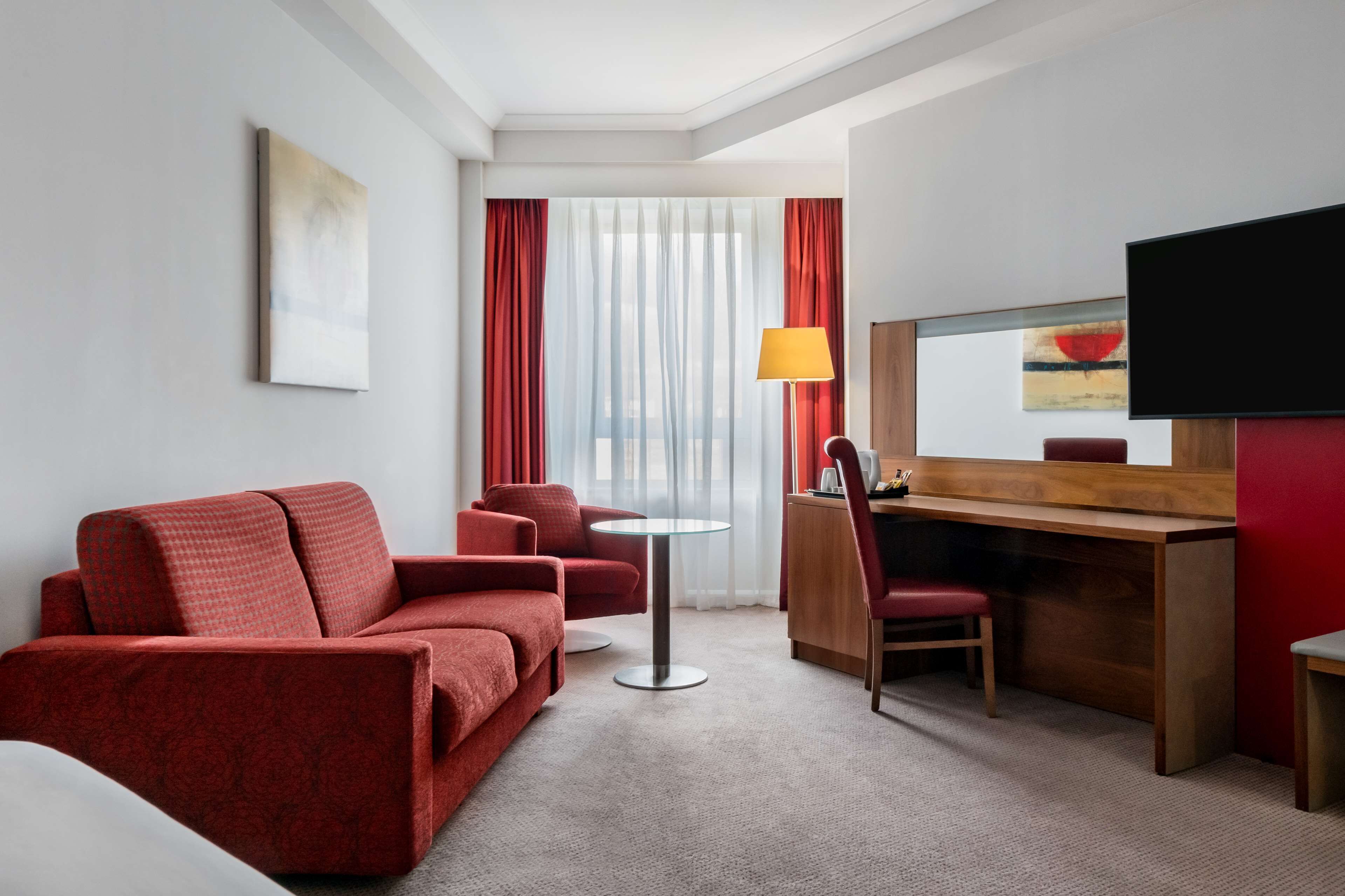 Une chambre d’hôtel avec un canapé rouge, une petite table ronde, un bureau avec une chaise, une télévision et une fenêtre avec des rideaux rouges.