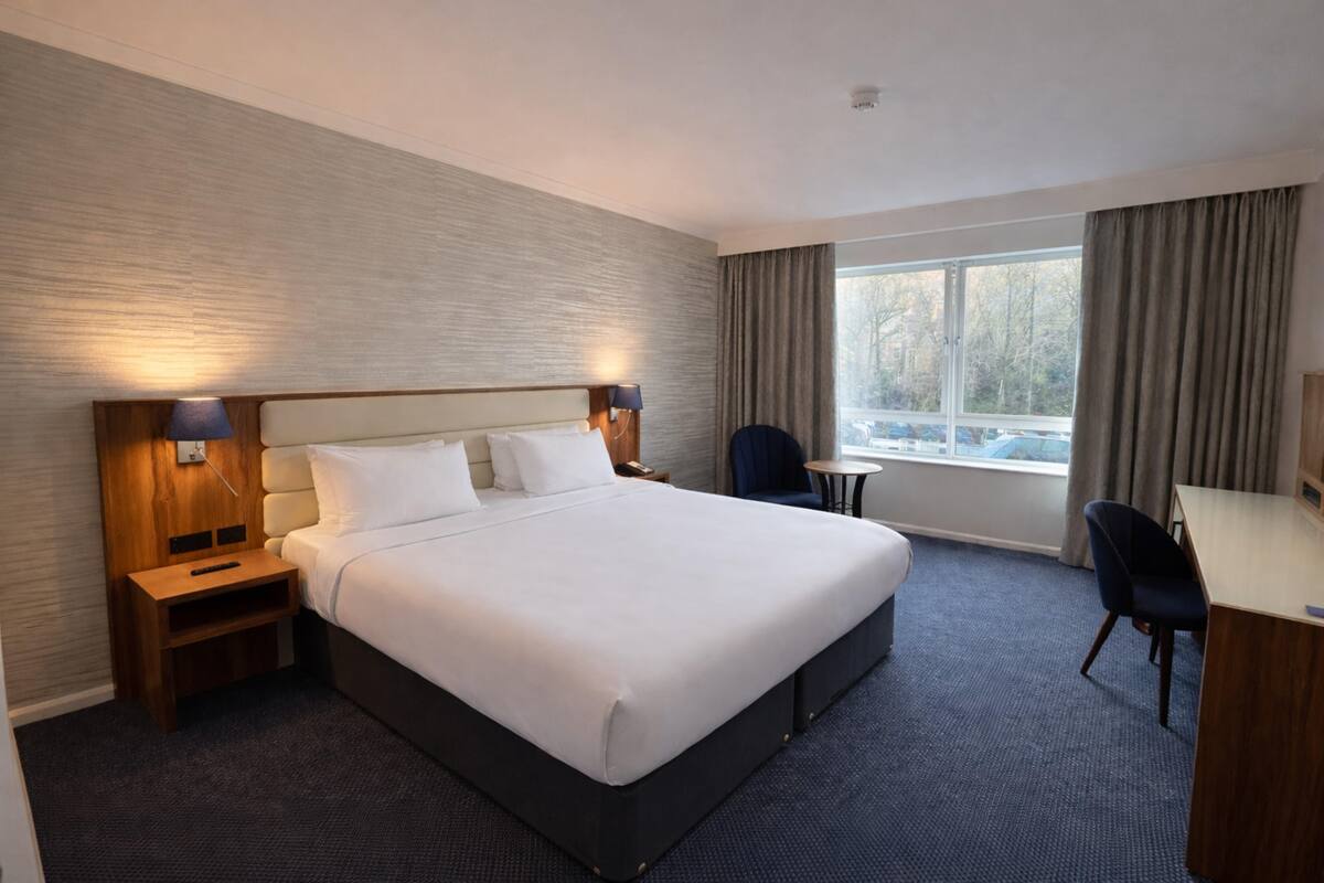 Une chambre d’hôtel avec un grand lit, un bureau, une chaise et une fenêtre avec des rideaux.