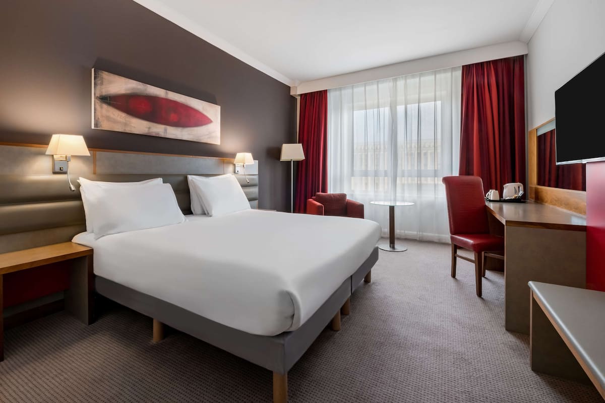 Une chambre d’hôtel avec un grand lit, deux tables de chevet avec des lampes, un bureau avec une chaise, une petite table avec une tasse et un livre, et une grande fenêtre avec des rideaux.