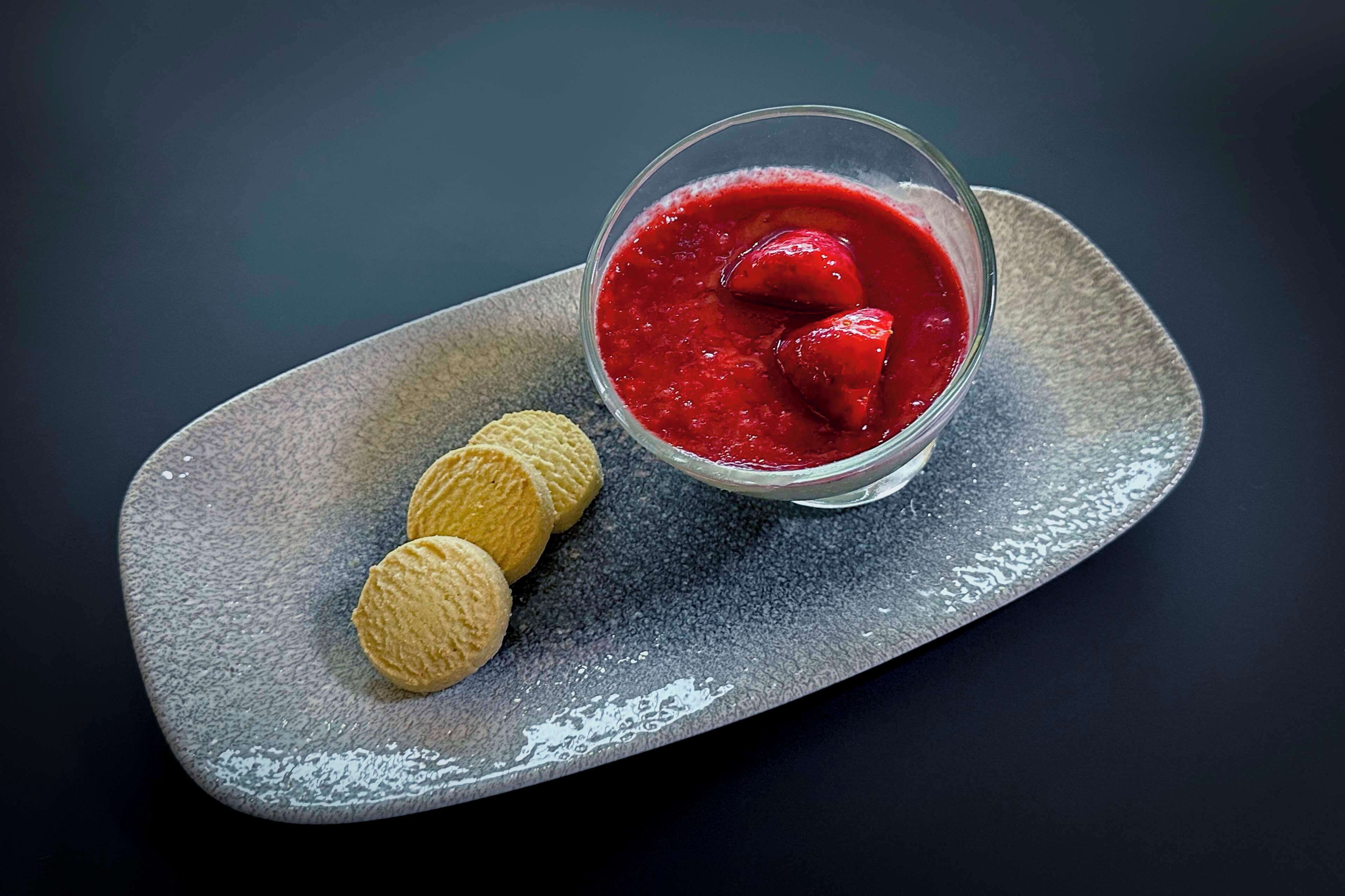 A dessert with a glass of red sauce and three cookies on a textured plate.