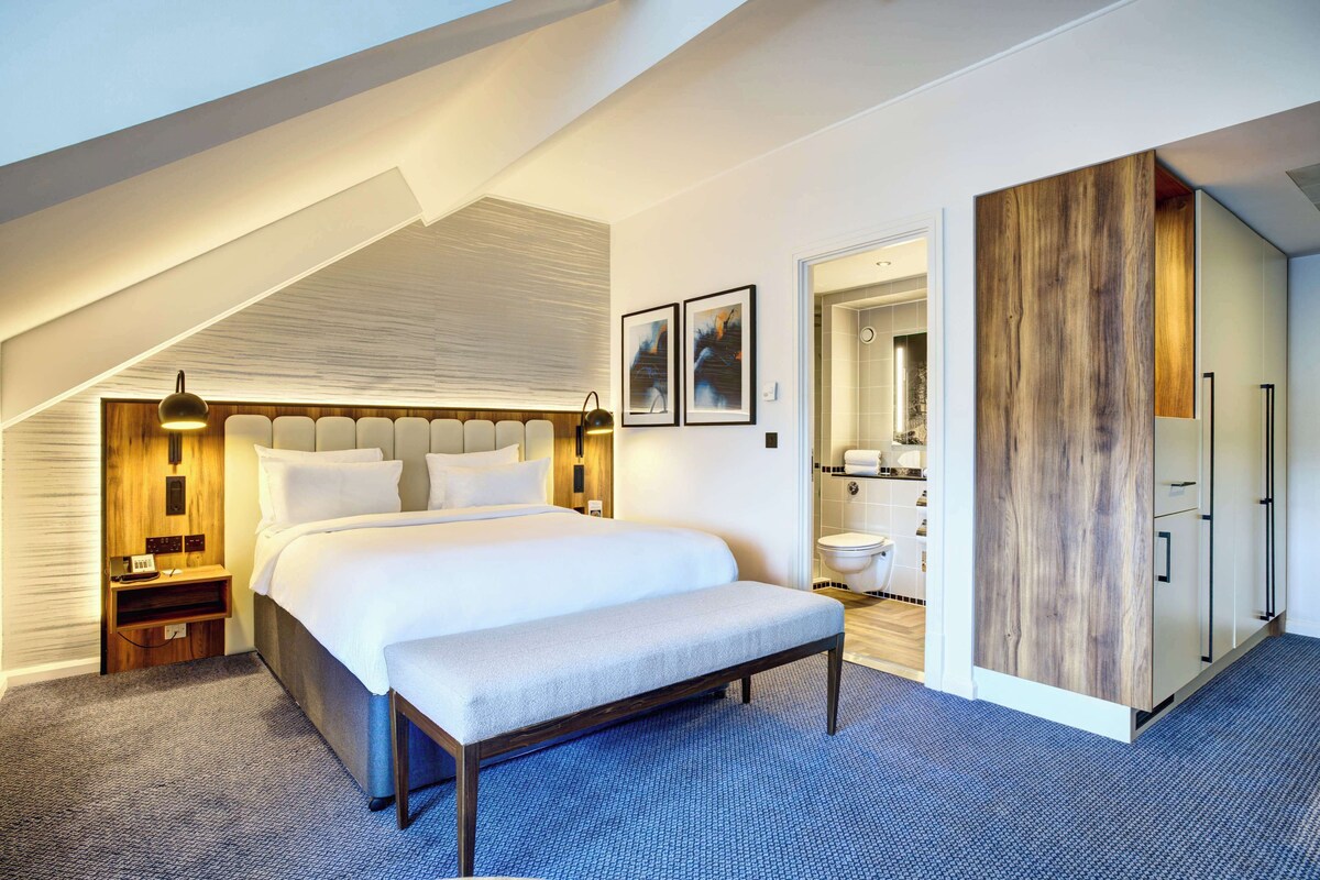 A modern hotel room with a large bed, a bench, a wooden wardrobe, and a bathroom visible through an open door.