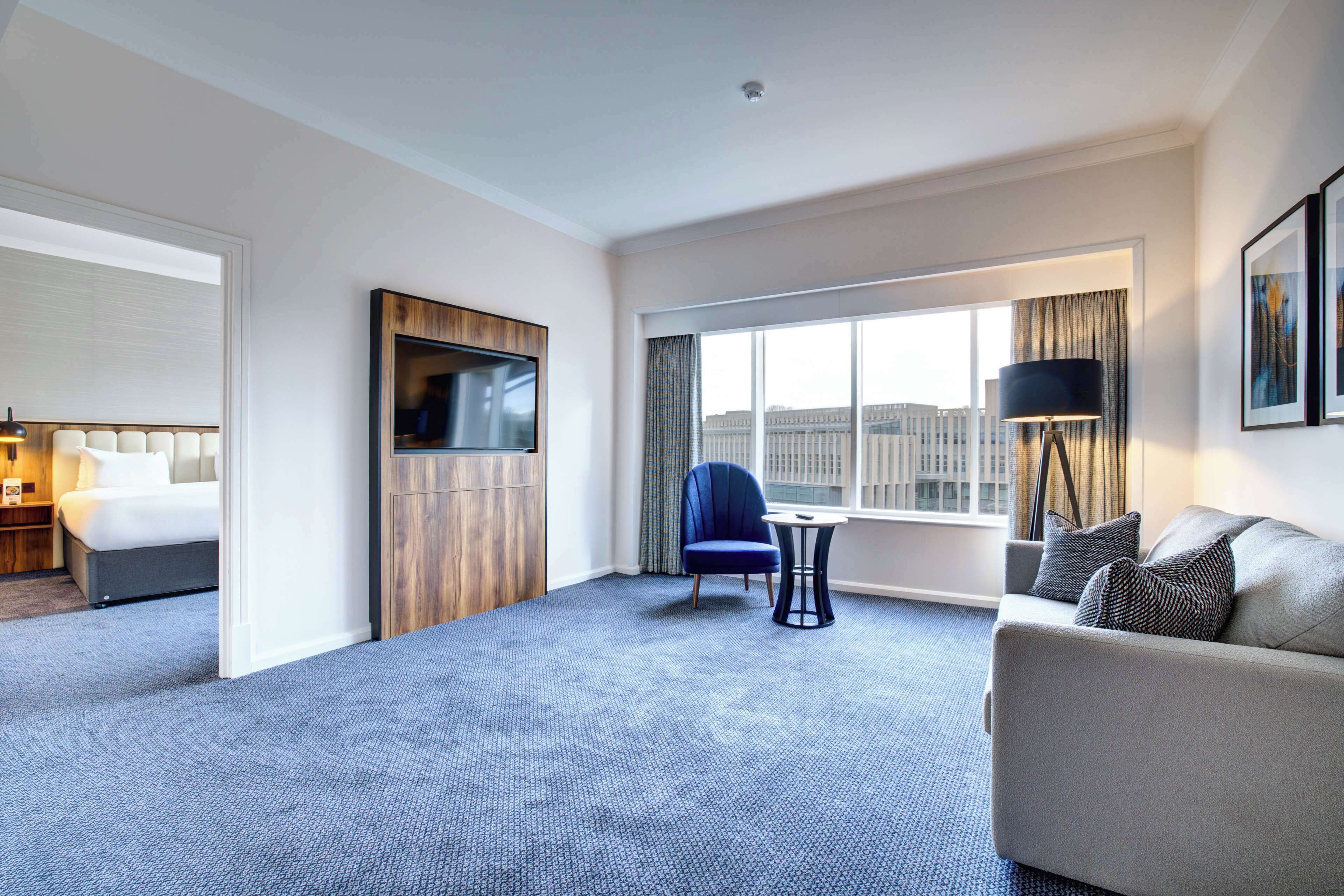 A hotel room with a bed, a sofa, a chair, a TV, and a large window with a city view.