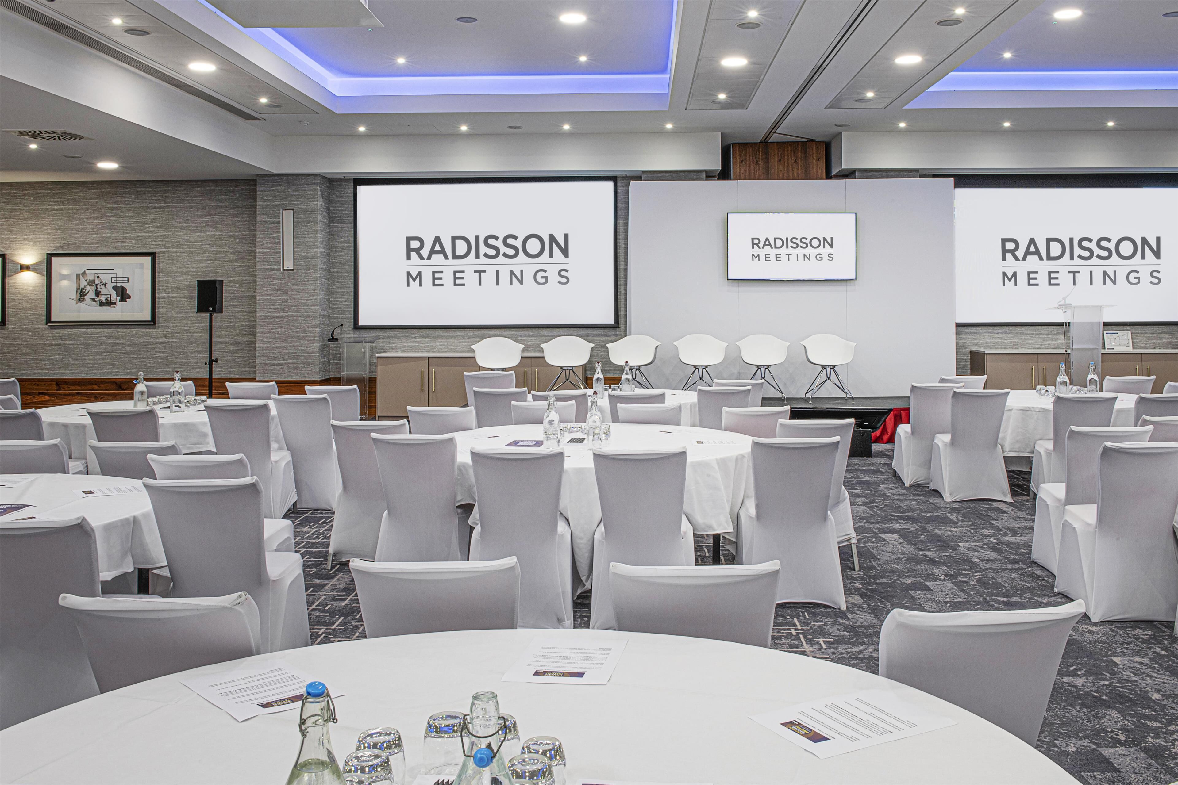A conference room with multiple tables and chairs, equipped with nameplates and water bottles, and a large screen displaying "RADISSON MEETINGS".