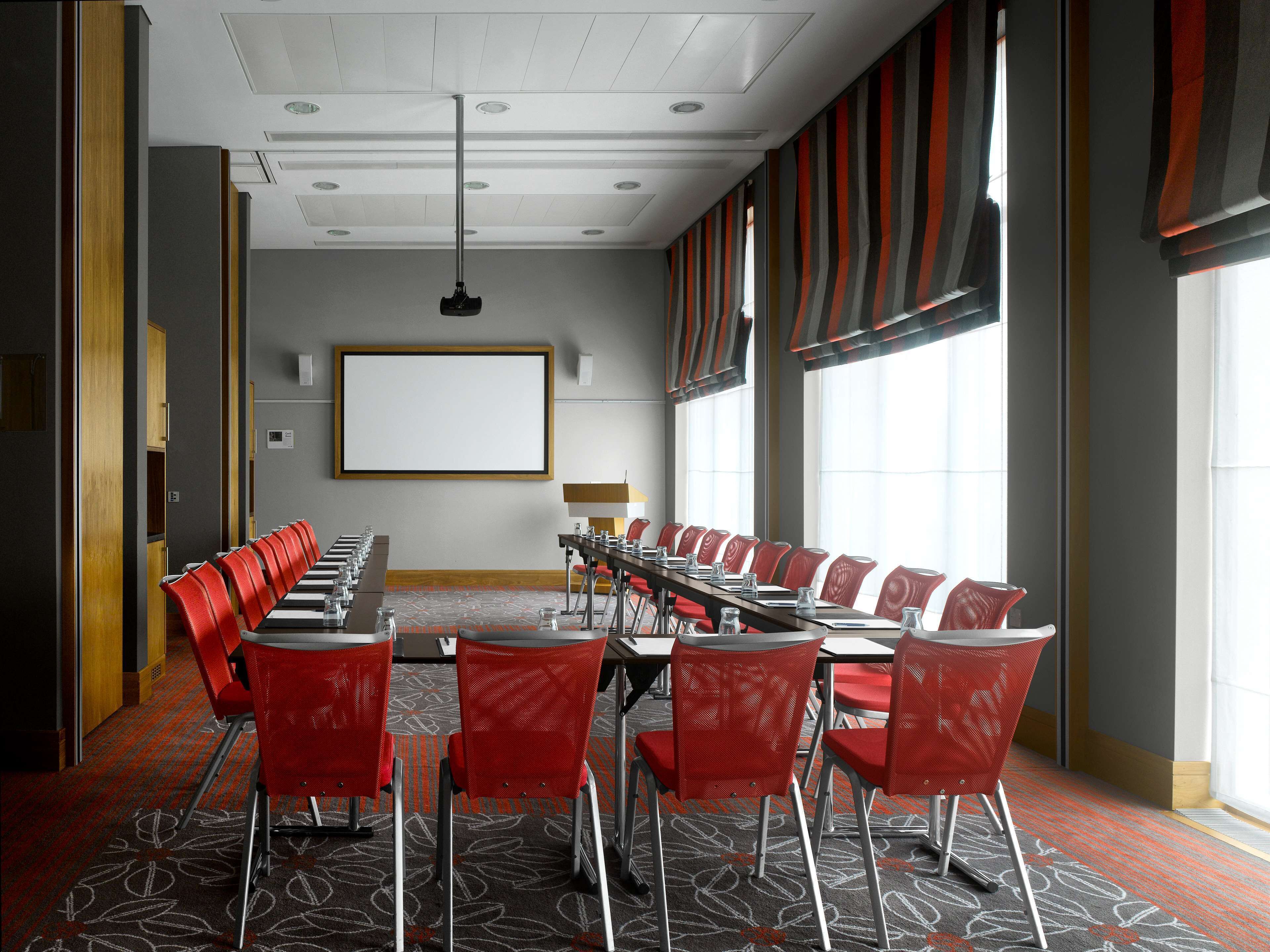 A conference room with a long table, red chairs, a projector screen, and large windows.