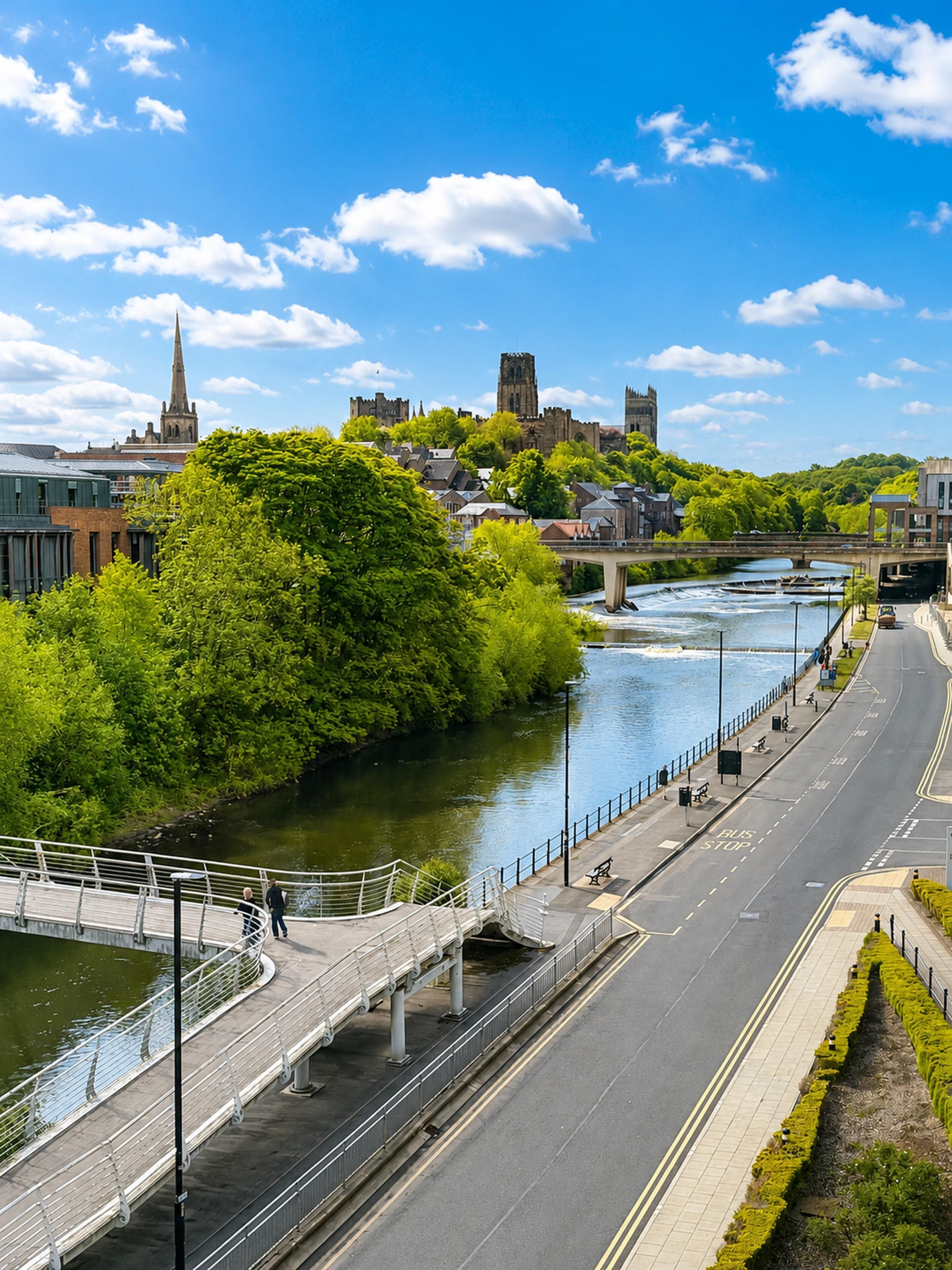 A riverside road with a pedestrian bridge, a river, and buildings in the background.