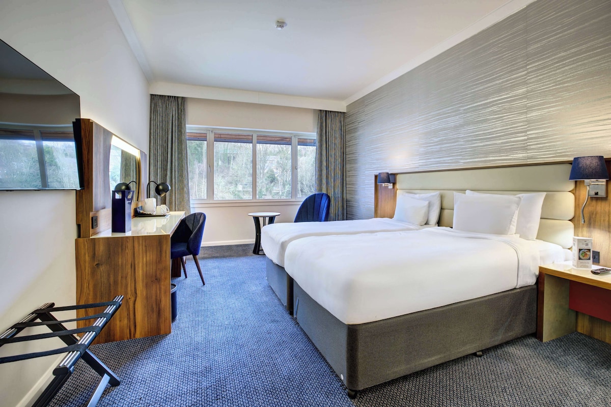 A hotel room with a large bed, a desk with a chair, a small table, and a window with curtains.