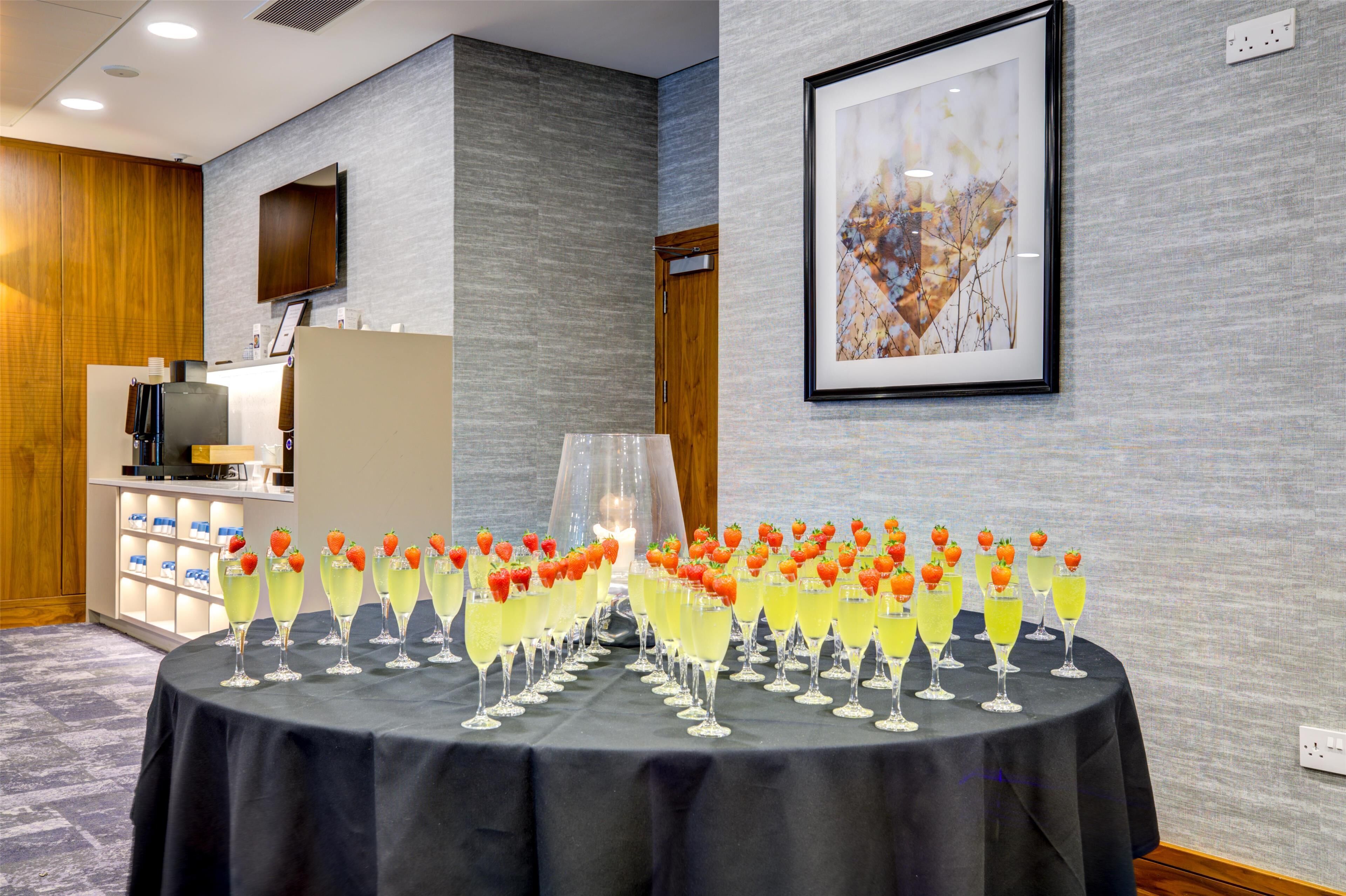 A table with multiple glasses of yellow cocktails garnished with strawberries, a wooden cabinet, and a framed abstract painting on the wall.