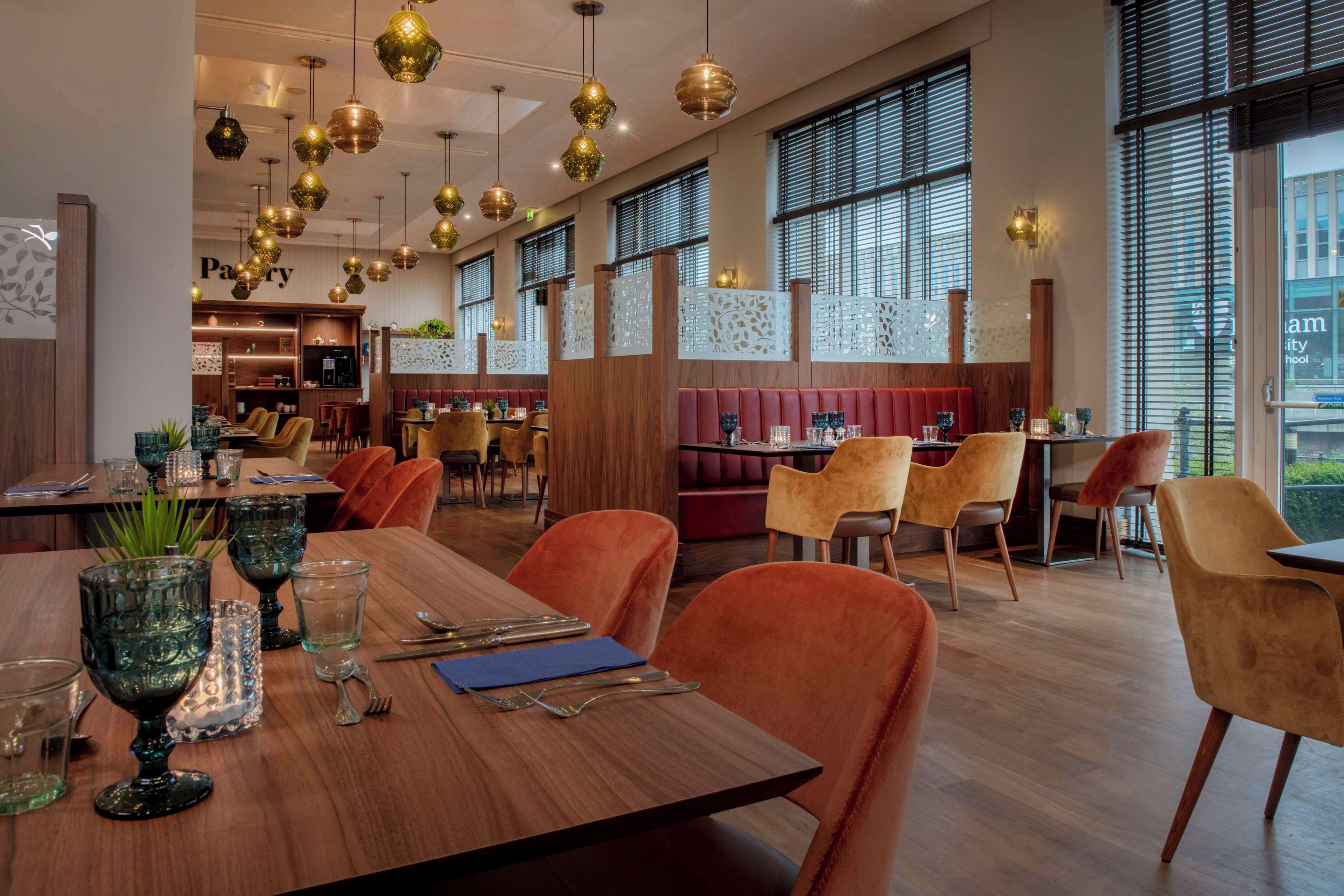 A restaurant with wooden tables and orange chairs, large windows, and hanging gold and black pendant lights.