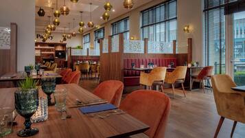 A restaurant with wooden tables and orange chairs, large windows, and hanging gold and black pendant lights.