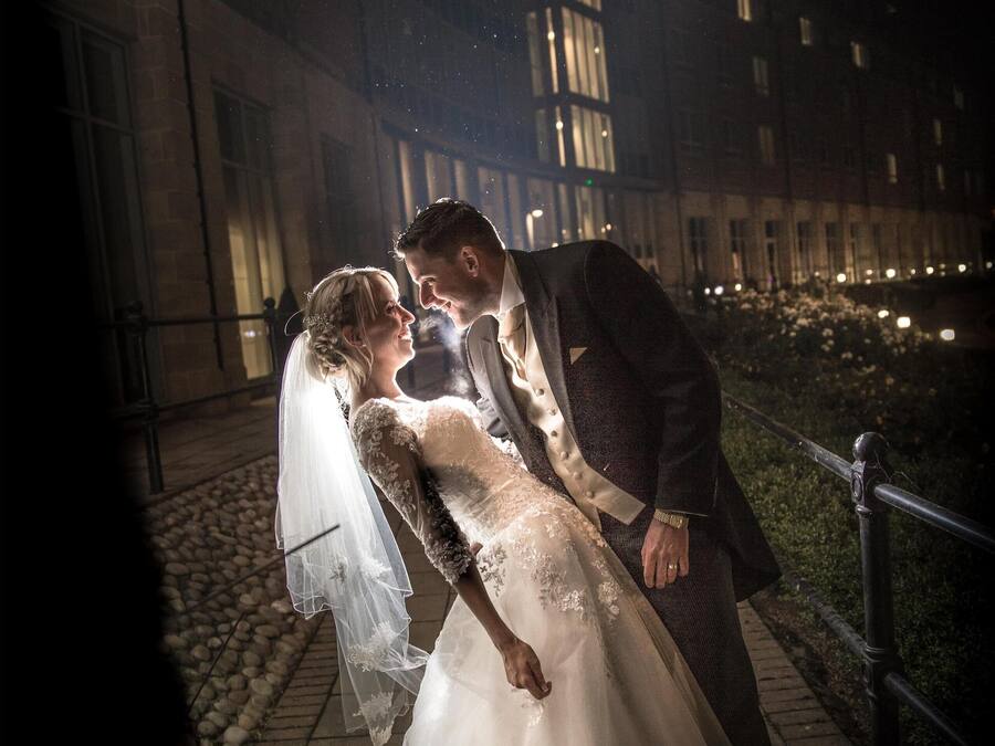 A couple embracing on a nighttime outdoor area with a building in the background.