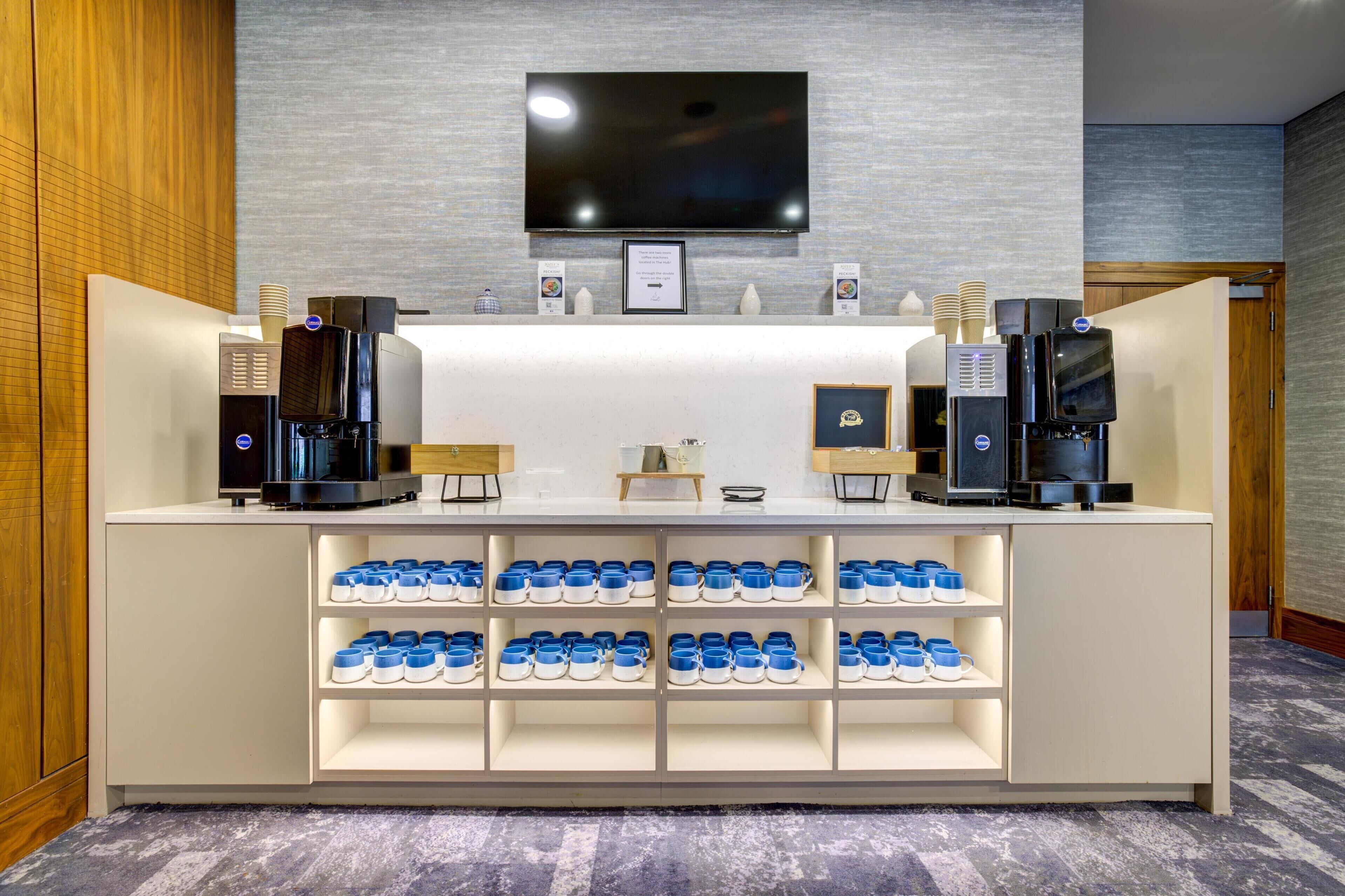 Zona de recepción de un hotel con un televisor de pantalla plana en la pared, una estación de café con varias cafeteras y una exhibición de tazas azules y blancas.