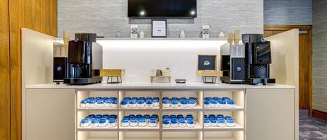 Zona de recepción de un hotel con un televisor de pantalla plana en la pared, una estación de café con varias cafeteras y una exhibición de tazas azules y blancas.