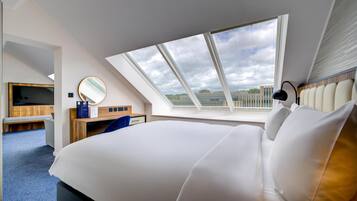 Quarto de hotel moderno com uma cama grande, escrivaninha, cadeira, televisão, espelho e claraboia.