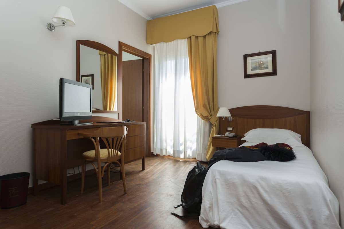 A hotel room with a wooden desk, a chair, a bed, a nightstand, a lamp, and a window with curtains.