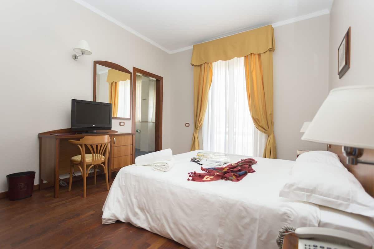 A hotel room with a bed, a desk with a computer, a chair, a telephone, and a window with curtains.