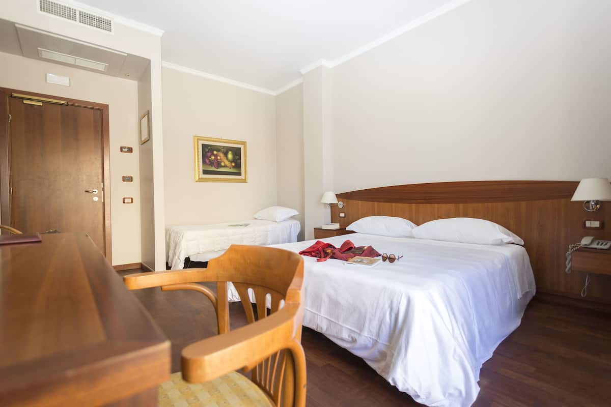 A hotel room with a wooden desk, two single beds, a wooden headboard, a bedside table with a lamp, and a framed picture on the wall.