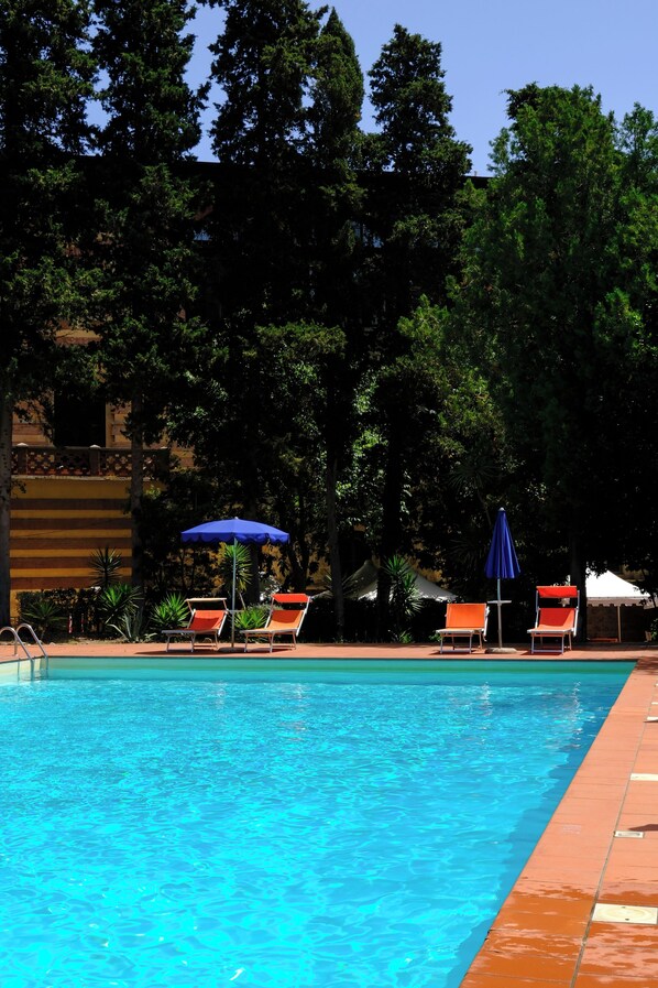 Una piscina con sillas de descanso y sombrillas, rodeada de árboles y un edificio al fondo.