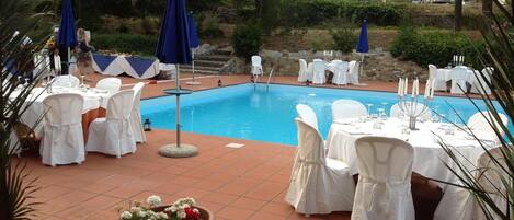 A poolside area with tables set for dining, surrounded by white chairs and blue umbrellas.