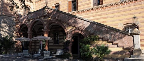 A courtyard with stone flooring, outdoor seating, and a building with arched windows and a staircase.