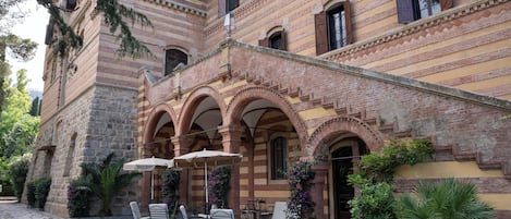Un patio con mobiliario exterior, suelo de piedra y un edificio con ventanas de arco y detalles de ladrillo.