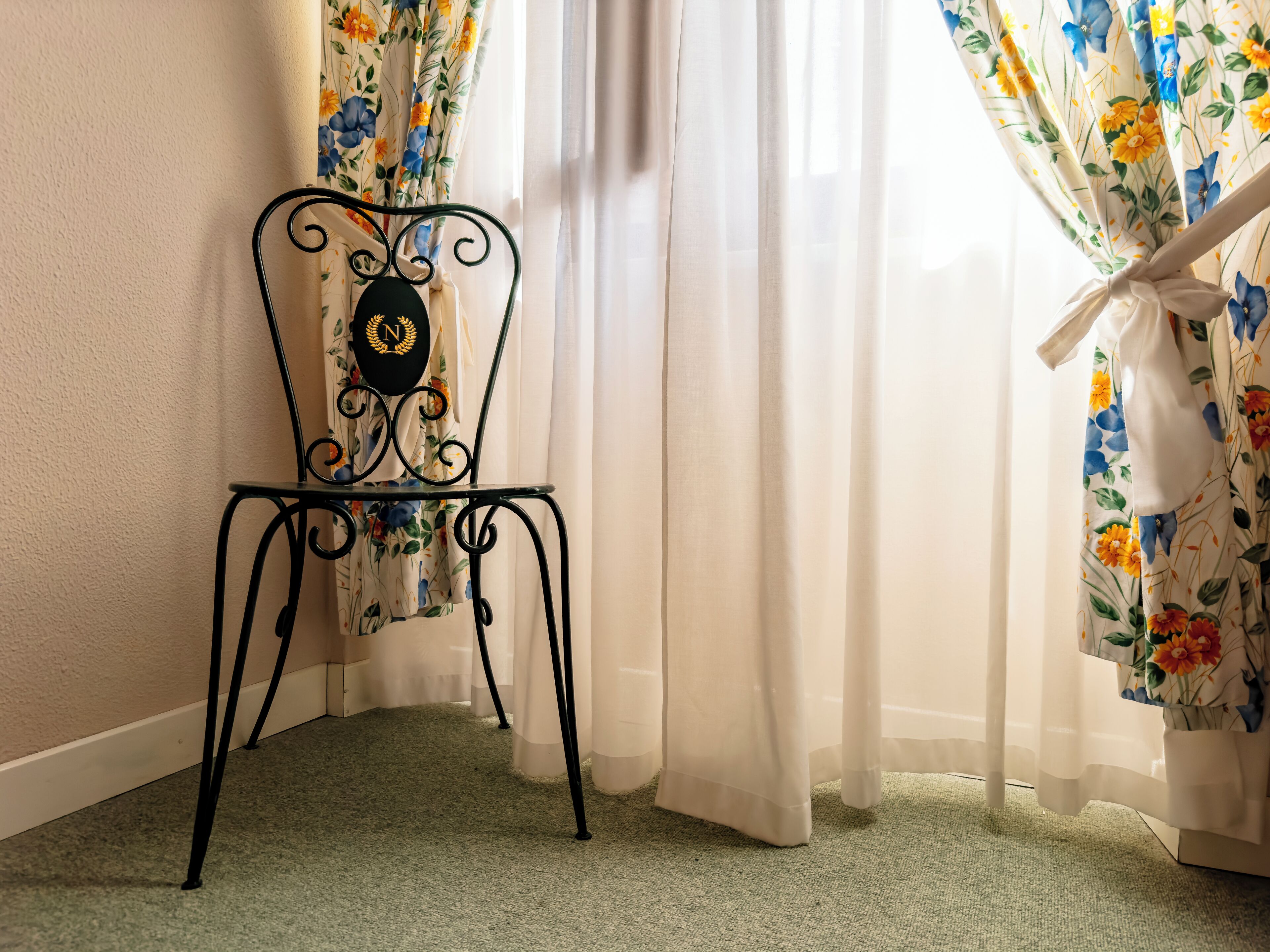 A corner of a room with a metal chair, a floral curtain, and a carpeted floor.