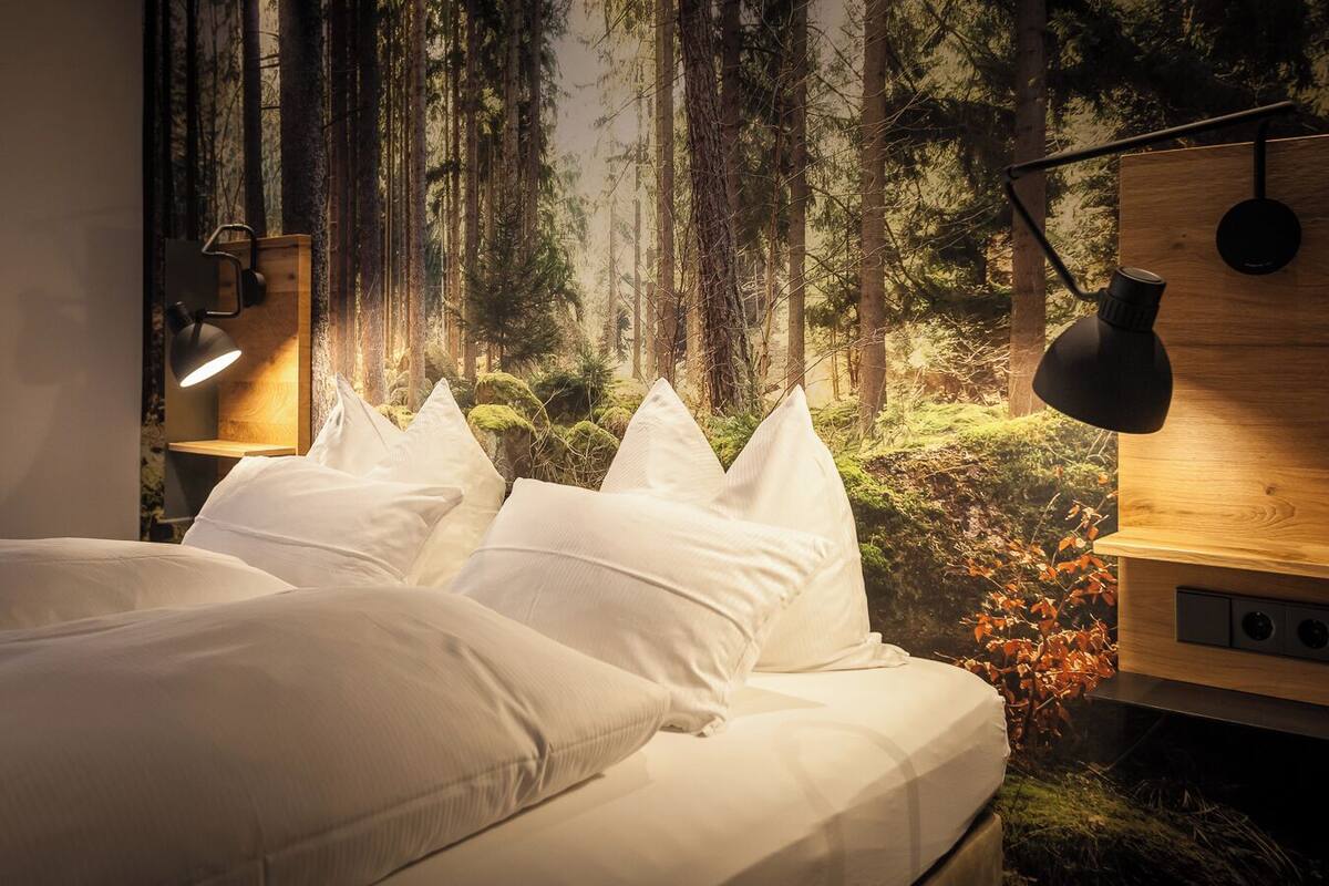 A bedroom with a bed, white pillows, a nightstand, a lamp, and a wall mural of a forest.