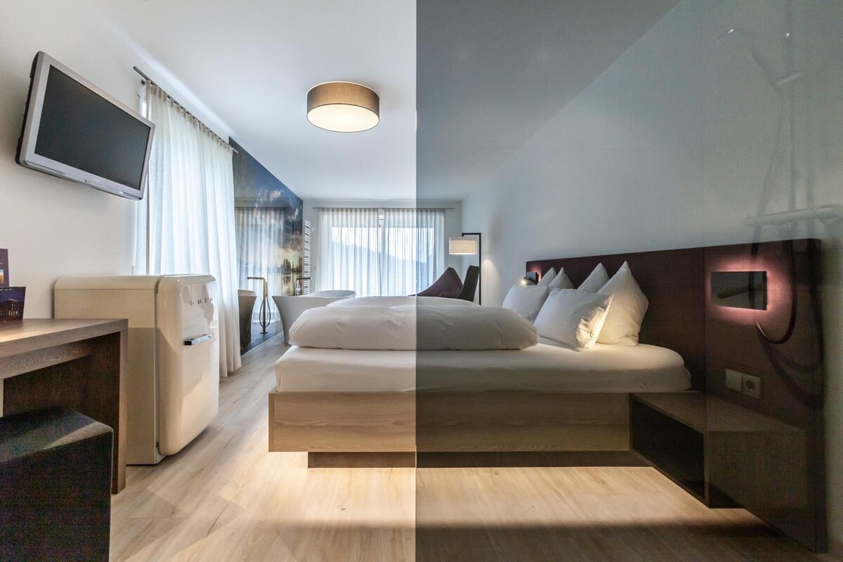 A hotel room with a bed, bedside table, TV, and a large window with curtains.