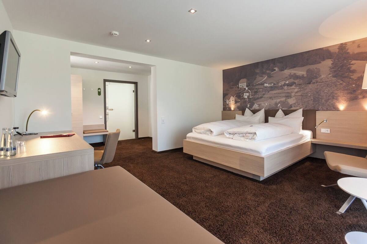 A modern hotel room with a large bed, a desk, a chair, and a painting on the wall.
