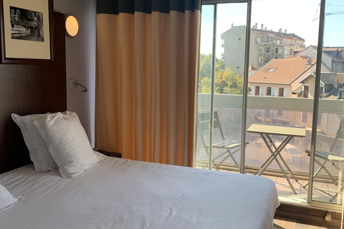 A hotel room with a large bed, a window with curtains, a framed picture on the wall, and a view of the city through the window.