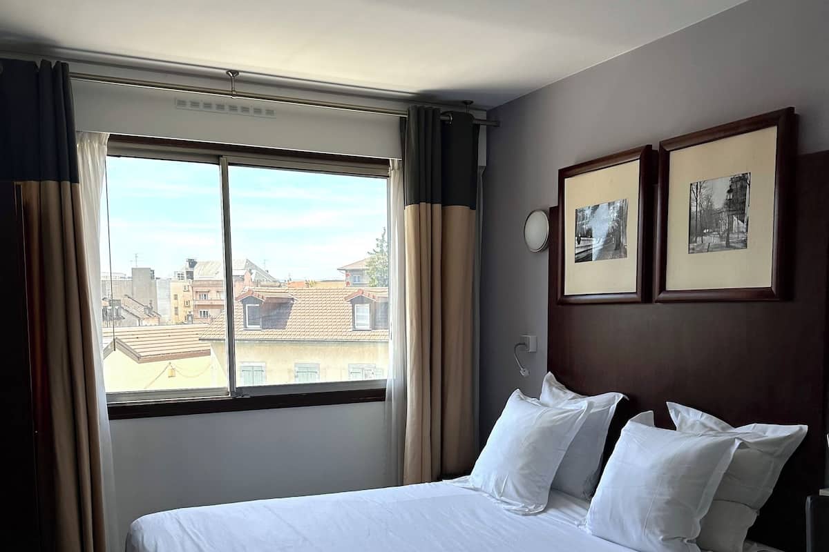 A hotel room with a large bed, a window with curtains, and two framed pictures on the wall.