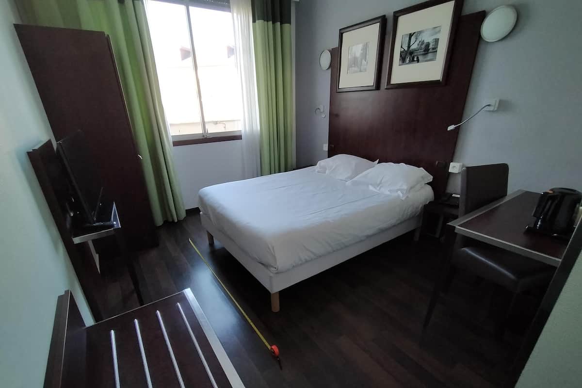 A hotel room with a bed, a desk with a chair, a coffee maker, a TV, and a window with curtains.