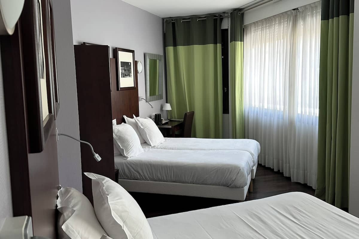 A hotel room with two beds, a desk, a chair, a lamp, and a window with curtains.