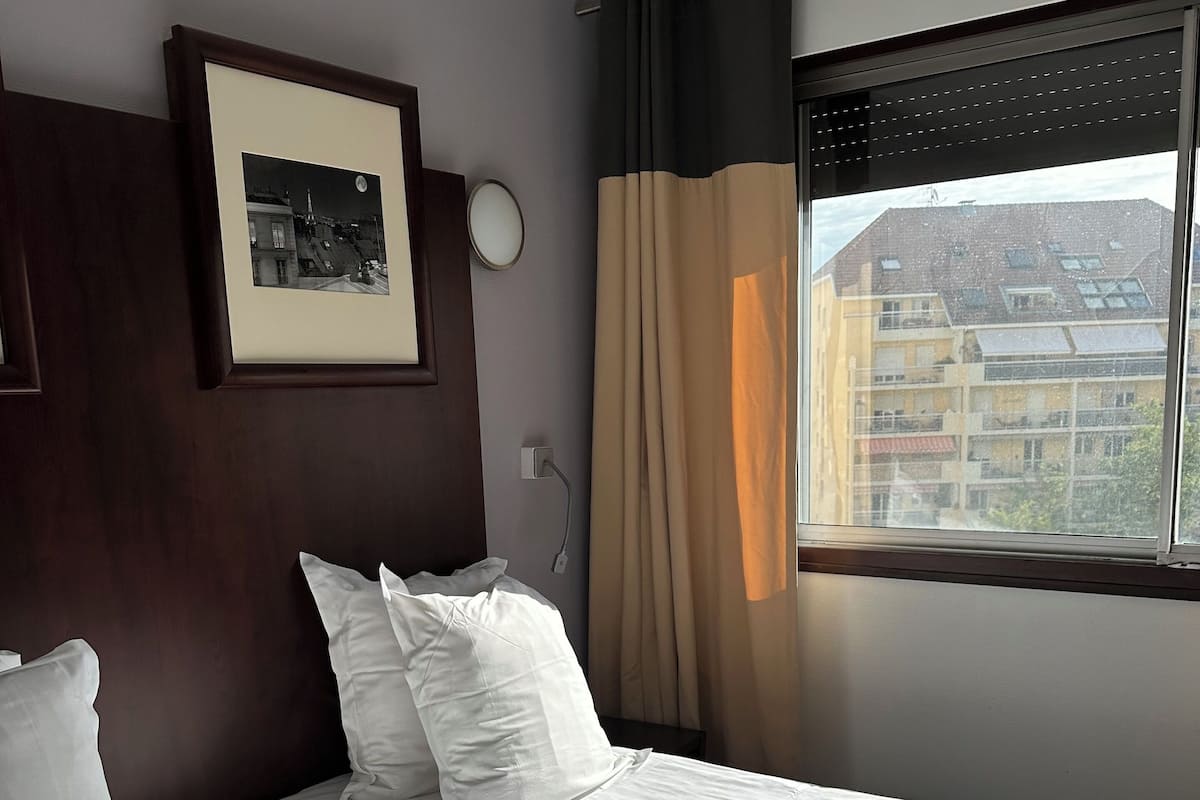 A hotel room with two beds, a framed picture on the wall, and a window with a view of buildings.