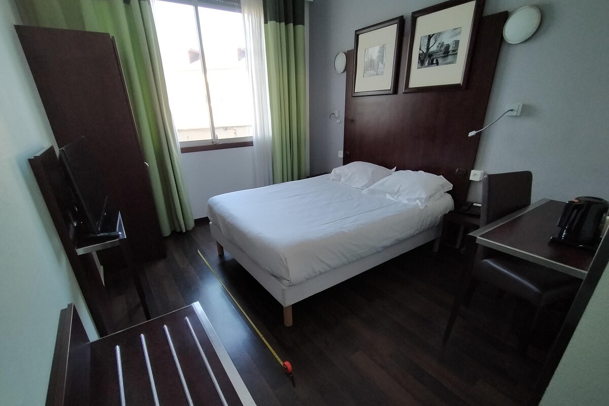 Una habitación de hotel con cama, escritorio con silla, cafetera, televisión y ventana con cortinas.
