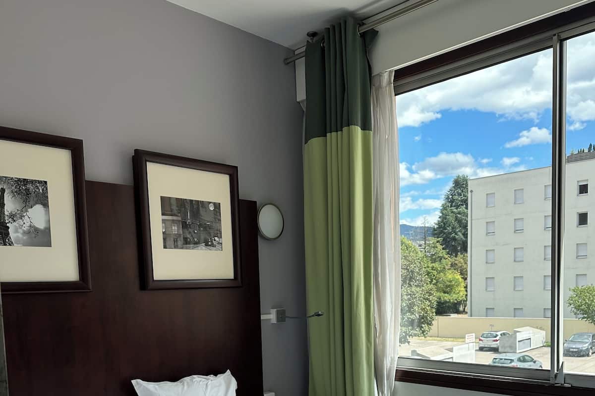 A hotel room with a bed, framed pictures on the wall, a window with curtains, and a view of buildings outside.
